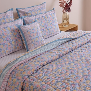 May include: A bed set featuring a floral pattern in shades of blue, pink, and yellow. The set includes a quilted comforter and several pillowcases. The bedding has a decorative border and is arranged on a bed with a neutral background.