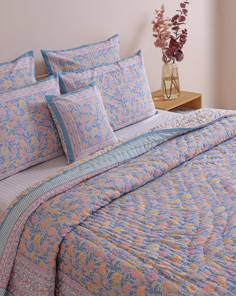 May include: A bed set featuring a floral pattern in shades of blue, pink, and yellow. The set includes a quilted comforter and several pillowcases. The bedding has a decorative border and is arranged on a bed with a neutral background.