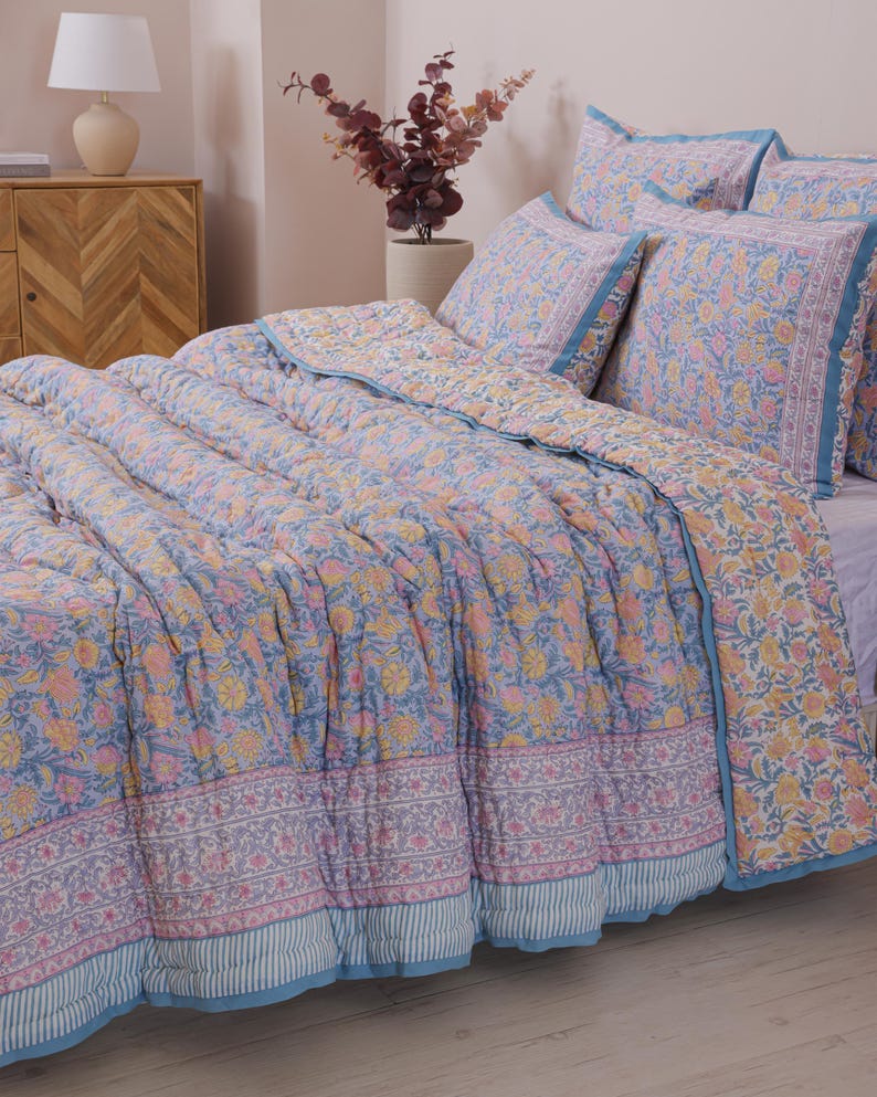 May include: A floral patterned quilt and pillow set in shades of blue, pink, and yellow. The bedding features a repeating flower design and a blue border. The quilt is on a bed with matching pillows, creating a coordinated bedroom set.