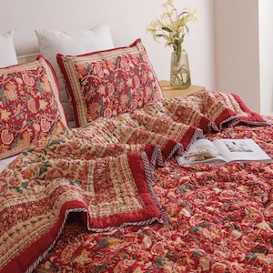 Pode incluir: Uma colcha acolchoada e duas fronhas retangulares a condizer com uma base vermelha vibrante e um padrão floral intrincado em tons de amarelo, laranja e verde. A roupa de cama apresenta uma borda decorativa e um acabamento às riscas.