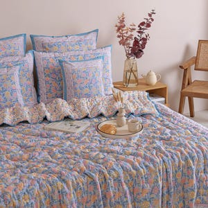 May include: A bed is covered with a blue, pink, and yellow floral patterned quilt and matching pillows. The pillows have a blue border. A tray with a croissant and coffee sits on the quilt. A vase of flowers and a teapot are on a wooden side table.
