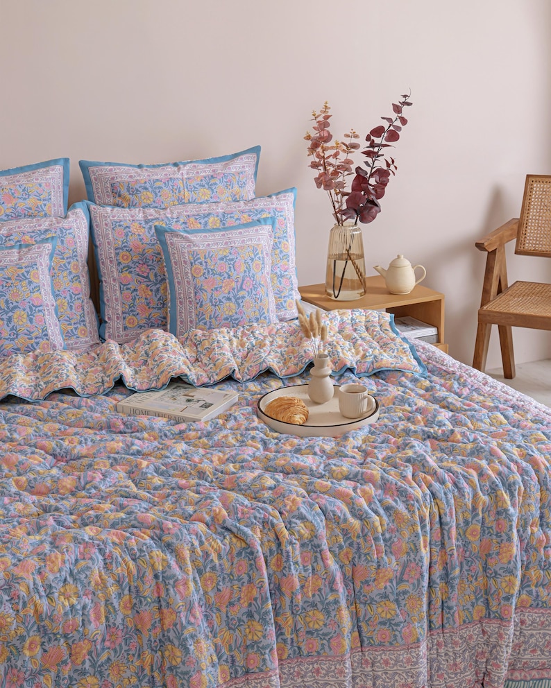 May include: A bed is covered with a blue, pink, and yellow floral patterned quilt and matching pillows. The pillows have a blue border. A tray with a croissant and coffee sits on the quilt. A vase of flowers and a teapot are on a wooden side table.