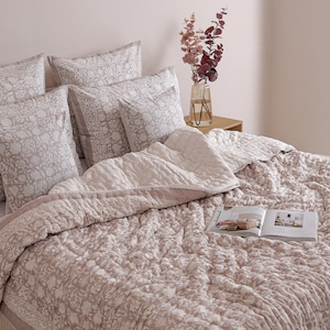 May include: A bed with a floral patterned quilt and matching pillowcases in shades of pink and white. A striped blanket and a book are on the bed. A vase of flowers sits on a wooden nightstand.