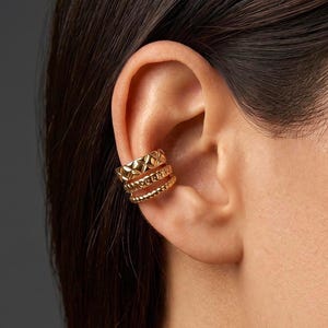 May include: A gold-toned ear cuff featuring three distinct bands. The top band has a textured diamond-shaped pattern, while the middle and bottom bands consist of a smooth, rounded wire design. The piece sits securely on the outer ear cartilage.