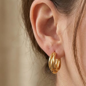 May include: Close-up of a gold hoop earring with a multi-layered design. The earring is worn on an ear, with the focus on the shiny, metallic texture and the way the light reflects off the curved surfaces. The earrings are a classic jewelry item.