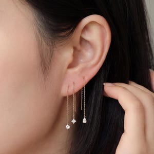 Puede incluir: Pendientes colgantes dorados. Un par presenta una gema cuadrada, otro una gema en forma de lágrima y el tercero es una barra simple. Los pendientes cuelgan de la oreja.