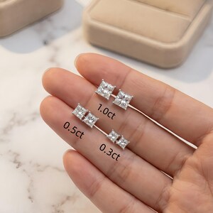 Puede incluir: Tres pares de pendientes de diamantes de talla cuadrada en diferentes tama&ntilde;os: 0,3 ct, 0,5 ct y 1,0 ct. Los pendientes se muestran en finos postes plateados. La imagen est&aacute; sobre un fondo neutro, con una caja de joyer&iacute;a visible.