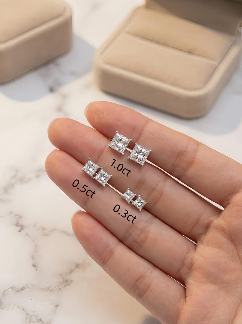Puede incluir: Tres pares de pendientes de diamantes de talla cuadrada en diferentes tama&ntilde;os: 0,3 ct, 0,5 ct y 1,0 ct. Los pendientes se muestran en finos postes plateados. La imagen est&aacute; sobre un fondo neutro, con una caja de joyer&iacute;a visible.