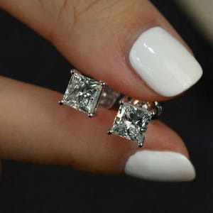 Puede incluir: Un par de pendientes de diamantes de talla cuadrada. Los diamantes est&aacute;n engastados en una montura de plata u oro blanco. Los pendientes est&aacute;n sostenidos por una persona con esmalte de u&ntilde;as blanco.