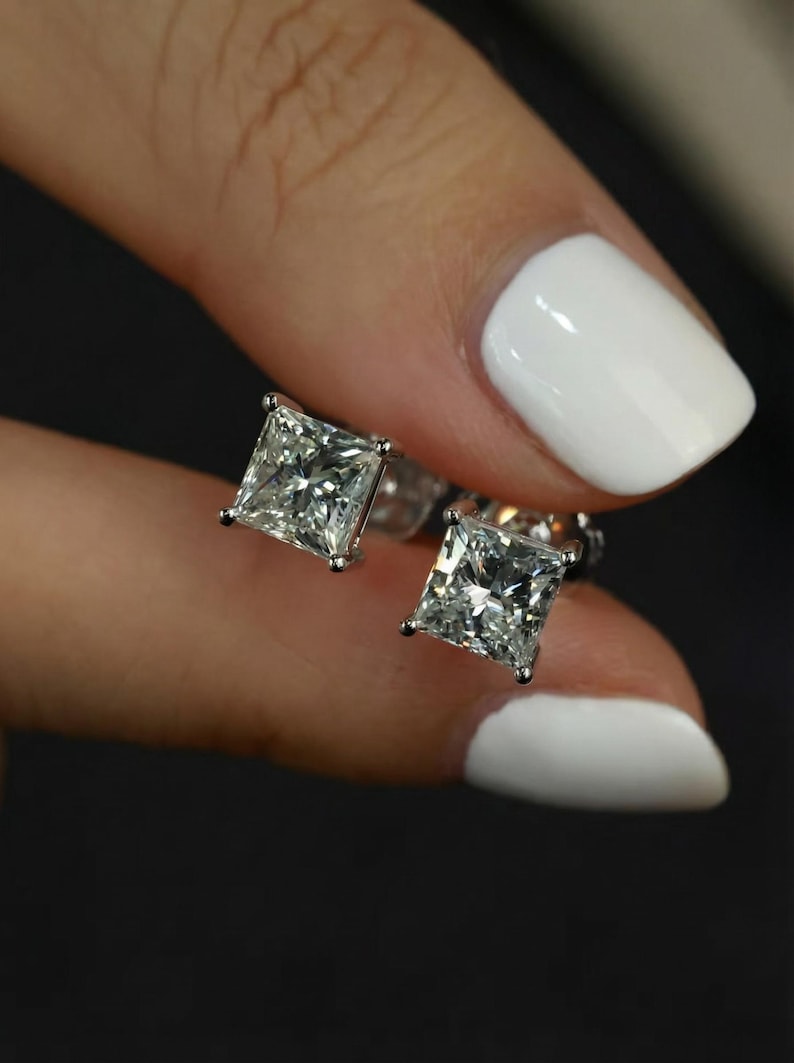 Puede incluir: Un par de pendientes de diamantes de talla cuadrada. Los diamantes est&aacute;n engastados en una montura de plata u oro blanco. Los pendientes est&aacute;n sostenidos por una persona con esmalte de u&ntilde;as blanco.