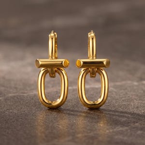 Könnte beinhalten: Ein Paar polierte goldfarbene Creolen mit vertikalem Design, bestehend aus einem glatten, abgerundeten Stab, der mit einem hängenden ovalen Glied verbunden ist. Die Ohrringe haben eine reflektierende Metalloberfläche und einen Klappverschluss.