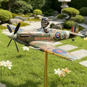 Può includere: Un modello di aereo con un design mimetico, con il logo degli Iron Maiden e Eddie the Head come pilota. L'aereo ha un'elica ed è montato su un supporto dorato. Il testo "IRON MAIDEN" e "1975-2025 Anniversary" sono visibili.