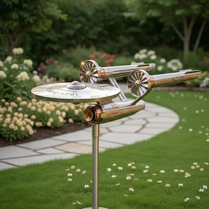 Puede incluir: Una escultura metálica de la nave estelar Enterprise, con un casco principal plateado en forma de disco y dos góndolas de motor. La escultura está montada sobre un poste plateado y colocada en un jardín cubierto de hierba con un camino de piedra y plantas florecientes.