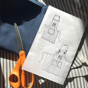 Puede incluir: Un boceto de un delantal con la letra "M" en papel blanco, junto a una tela azul marino y unas tijeras naranjas. El diseño del delantal presenta un patrón de rayas y bolsillos. El fondo es una tela a rayas.