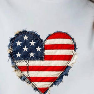 Op de afbeelding: Een lichtblauw t-shirt met een hartvormige afbeelding op de borst. Het hartontwerp bevat het patroon van de Amerikaanse vlag met rode en witte horizontale strepen en een blauw gedeelte met witte sterren, afgewerkt met gerafelde randen.