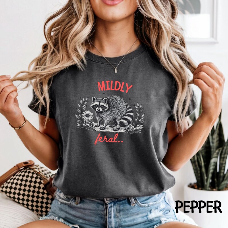 May include: A charcoal gray crew neck t-shirt featuring a black and white illustration of a raccoon surrounded by foliage and a flower. The text "MILDLY" appears in red above the raccoon, and the word "feral.." is printed in red below it.
