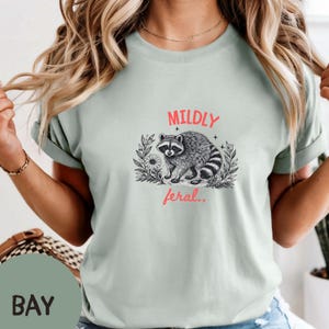 May include: A light sage green t-shirt featuring a black and white line art illustration of a raccoon surrounded by foliage and flowers. The text MILDLY is printed in red above the raccoon, and the text feral.. is printed in red below it.