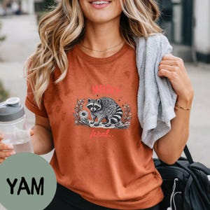 May include: A rust-colored short-sleeve t-shirt featuring a black and white graphic of a raccoon surrounded by foliage. The text "MILDLY" appears in red above the raccoon, and the word "feral.." is printed in red below the illustration.