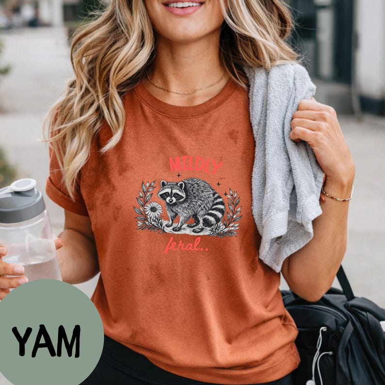 May include: A rust-colored short-sleeve t-shirt featuring a black and white graphic of a raccoon surrounded by foliage. The text "MILDLY" appears in red above the raccoon, and the word "feral.." is printed in red below the illustration.