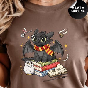 May include: A brown t-shirt featuring a graphic of a black dragon wearing round glasses and a red and yellow striped scarf. The dragon sits on a stack of books next to a white owl, a wand, and a golden winged ball, surrounded by small yellow stars.