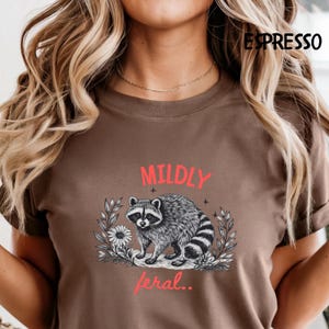 May include: An espresso brown t-shirt featuring a black and white illustration of a raccoon surrounded by leaves and a flower. The text MILDLY is printed in red above the raccoon, and the text feral.. is printed in red below the raccoon.