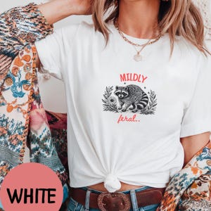 May include: A white short-sleeved t-shirt featuring a black and white graphic illustration of a raccoon surrounded by foliage. The text MILDLY is printed in red above the raccoon, and the text feral.. is printed in red below the raccoon.
