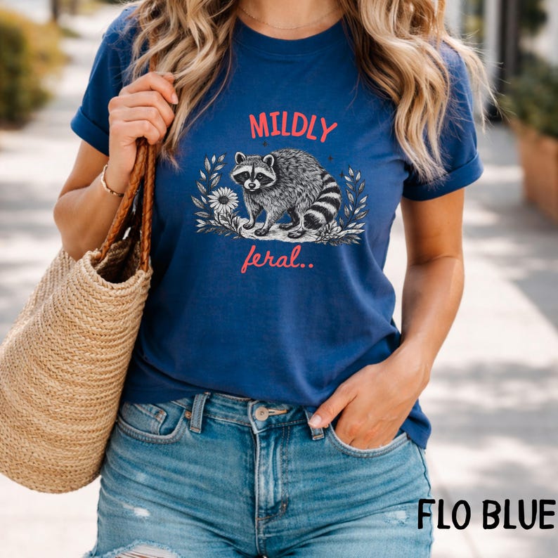 May include: A blue t-shirt featuring a graphic of a raccoon surrounded by floral elements. The text MILDLY is printed in red above the raccoon, and the text feral.. is printed in red below the raccoon. The shirt has short sleeves and a crew neckline.