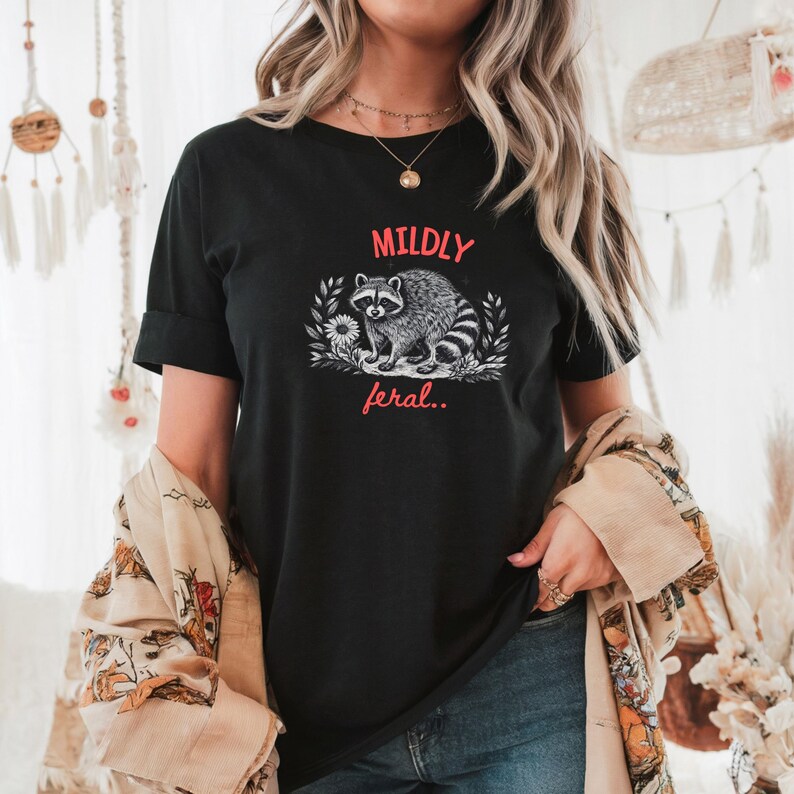 May include: A black short-sleeved t-shirt featuring a graphic illustration of a raccoon surrounded by foliage. The text MILDLY is printed in red above the raccoon, and the text feral.. is printed in red below the raccoon in a script font.