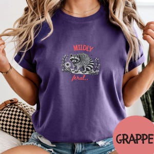 May include: A purple short-sleeve t-shirt featuring a graphic of a raccoon surrounded by foliage. The text "MILDLY" appears in coral above the raccoon, and the text "feral.." appears in coral below the raccoon on the center of the chest.