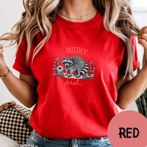 May include: A bright red short-sleeved t-shirt featuring a black and white graphic of a raccoon surrounded by leaves and a flower. The text MILDLY is printed above the raccoon and the text feral.. is printed below the raccoon in a light font.