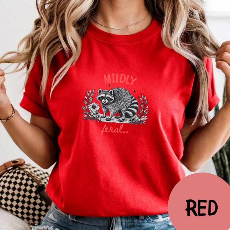 May include: A bright red short-sleeved t-shirt featuring a black and white graphic of a raccoon surrounded by leaves and a flower. The text MILDLY is printed above the raccoon and the text feral.. is printed below the raccoon in a light font.