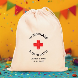 May include: A cream-colored drawstring pouch featuring a red cross graphic and black text that reads IN SICKNESS & IN HEALTH, JERRY & TOM, and 11.11.2026. The fabric bag has a gathered top closure with matching drawstrings on both sides.