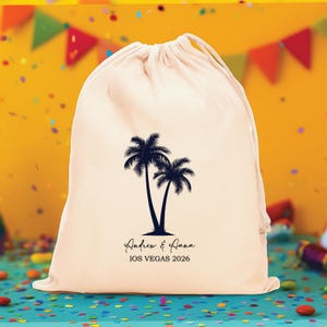 May include: A cream-coloured drawstring pouch featuring a black graphic of two palm trees. Below the trees, the text reads "Andrew & Aana" in a script font, followed by the text "IOS VEGAS 2026" in a simple, clean, sans-serif typeface.