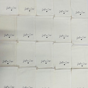 Puede incluir: Una colecci&oacute;n de veinte bolsas rectangulares de color blanco roto con cord&oacute;n, dispuestas en cuadr&iacute;cula. Cada bolsa presenta el texto impreso en negro Just in Case con un peque&ntilde;o s&iacute;mbolo de coraz&oacute;n centrado debajo de las letras.