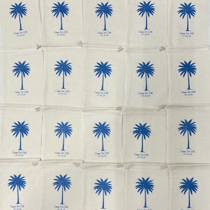 Puede incluir: Una colecci&oacute;n de veinte bolsas de tela color hueso con cord&oacute;n, dispuestas en cuadr&iacute;cula. Cada bolsa presenta un gr&aacute;fico de palmera azul impreso sobre el texto Casa De DR y la fecha 05.16.26. La tela tiene una textura tejida visible.