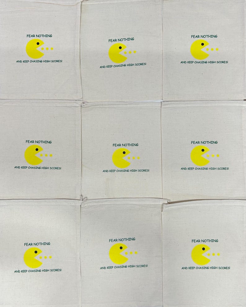 Puede incluir: Nueve bolsas de algod&oacute;n de color blanco roto con cord&oacute;n dispuestas en una cuadr&iacute;cula. Cada bolsa presenta un gr&aacute;fico de personaje circular amarillo con un ojo negro y la boca abierta, junto al texto FEAR NOTHING AND KEEP CHASING HIGH SCORES en negro.