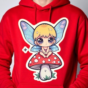 Könnte beinhalten: Roter Kapuzenpullover mit einem Cartoon-Feen-Design. Die Fee hat blonde Haare, blaue Flügel und große Augen und sitzt auf einem roten Pilz mit weißen Punkten. Die Fee trägt ein rosa Outfit. Das Design befindet sich auf der Vorderseite.