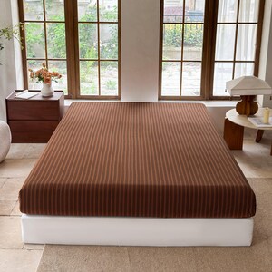 May include: A rectangular mattress featuring a brown fabric cover with a repeating pattern of thin, vertical black stripes. The mattress is positioned on a plain white base, presenting a clean, structured appearance with consistent, evenly spaced lines.