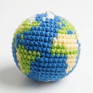 Puede incluir: Un globo terráqueo de ganchillo con océanos azules, continentes verdes y amarillos y regiones polares blancas. La esfera hecha a mano tiene un pequeño lazo de metal en la parte superior para colgar. El globo está sobre una superficie blanca.