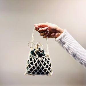 May include: A small, elegant handbag with a white pearl lattice design. The bag features a dark green velvet interior and a pearl handle. A gold-colored bow adorns the top. The bag is held by a person.