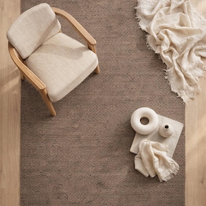 Puede incluir: Vista aérea de una ambientación de habitación con una alfombra rectangular marrón, un sillón beige con estructura de madera y objetos decorativos. La alfombra es texturizada. Un objeto blanco en forma de rosquilla y un jarrón pequeño están sobre un paño de lino.