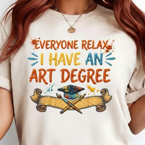 以下が含まれることがあります： 「EVERYONE RELAX I HAVE AN ART DEGREE」とカラフルな文字で書かれたベージュ色のTシャツ。デザインには、ペイントスプラッター、卒業帽子、筆、鉛筆が巻物の上に描かれています。