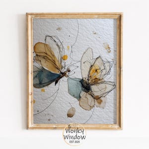 May include: A framed artwork featuring two abstract butterfly illustrations. The butterflies have tan, yellow, and blue watercolor wings with black line details. The background is a textured silver-gray. The frame is gold. The Wonky Window EST 2025 logo is in the bottom right.