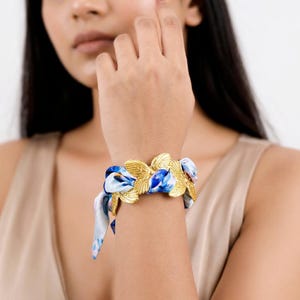 Könnte beinhalten: Ein goldfarbenes Metallarmband mit floralem Design. Das Armband ist mit blau-weißem Stoff verziert. Das Armband wird am Handgelenk getragen.