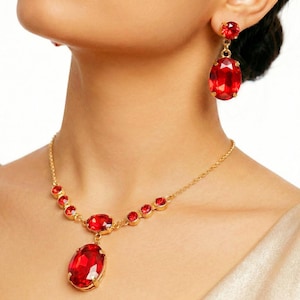 May include: A gold-toned necklace and earring set featuring large, oval, red gemstones. The necklace has a series of smaller red stones and a larger pendant. The earrings have a smaller stone at the top and a larger oval stone.