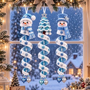 Könnte beinhalten: Drei hängende Weihnachtsdekorationen bestehend aus zwei gehäkelten Schneemännern und einem zentralen gehäkelten Weihnachtsbaum. Jedes Ornament hat ein langes, spiralförmiges weiß-blaues Band mit blauen Schneeflocken, das in zwei kleinen Metallkugeln endet.