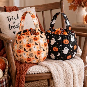 May include: Two fabric Halloween candy buckets with handles. One bucket is light orange with a pattern of ghosts, pumpkins, spiders, and webs. The other bucket is black with a pattern of white ghosts, orange pumpkins, and crescent moons.
