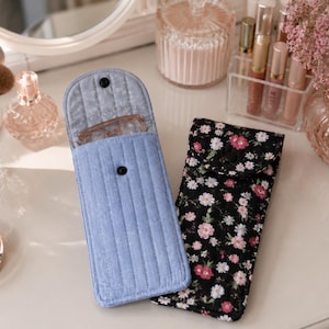 May include: Two rectangular fabric cases for eyeglasses, one in solid light blue with vertical quilting and a snap closure, and the other in black with a pink and white floral pattern, also featuring a snap closure and a flap design.