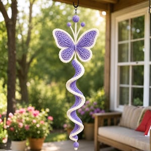 May include: A hanging crochet decoration featuring a purple and white butterfly at the top, connected to a long, spiraling purple and white tail. The piece is finished with small crocheted spheres at the top and bottom, suspended by a black hook.