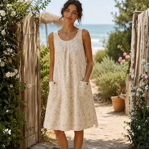 May include: A sleeveless knee-length dress in a light cream color featuring a delicate floral pattern in soft yellow and green tones. The dress has a round neckline with subtle pleating at the chest and two front patch pockets with small buttons.